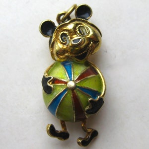 May include: A gold-toned metal charm featuring a cartoon character with black ears and a smiling face, holding a round ball decorated with segmented panels of translucent green, blue, and dark red enamel, finished with black enamel accents.