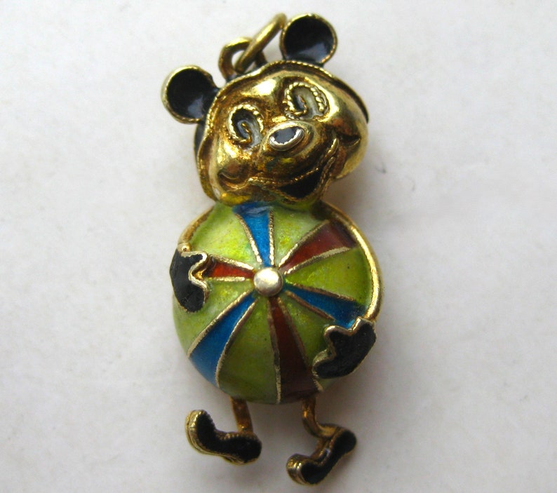May include: A gold-toned metal charm featuring a cartoon character with black ears and a smiling face, holding a round ball decorated with segmented panels of translucent green, blue, and dark red enamel, finished with black enamel accents.