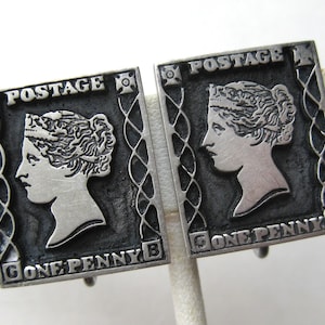 May include: A pair of silver-tone cufflinks. Each cufflink is rectangular and features a profile of a woman, with the words "POSTAGE" and "ONE PENNY B". The design has a dark background and a decorative border, reminiscent of a postage stamp.