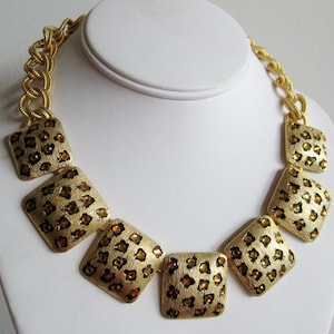 Op de afbeelding: Goudkleurige ketting met een ketting en vierkante hangers. De hangers hebben een textuur met een luipaardprint en amberkleurige accenten. De ketting wordt getoond op een witte buste.