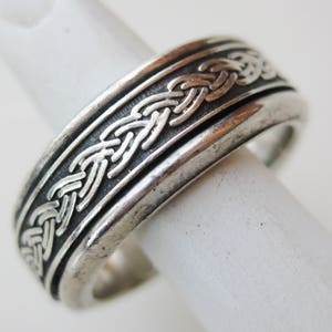 May include: A silver-toned ring with a Celtic knot design. The ring features a band with raised, interwoven patterns, and is displayed on a white surface. The ring's design is detailed and intricate, showcasing a classic style.