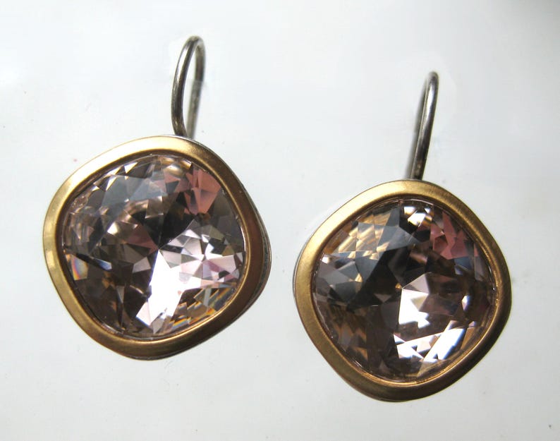 May include: A pair of earrings featuring square-shaped, faceted light pink crystals set in smooth, matte gold-toned metal bezels. Each earring is attached to a simple, curved silver-toned metal ear wire hook for hanging, showcasing a clean design.