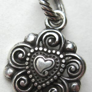 May include: A silver pendant featuring a central heart shape surrounded by intricate swirling metal patterns and a beaded border. The pendant is attached to a twisted rope-style silver bail at the top, showcasing a polished and oxidized finish.