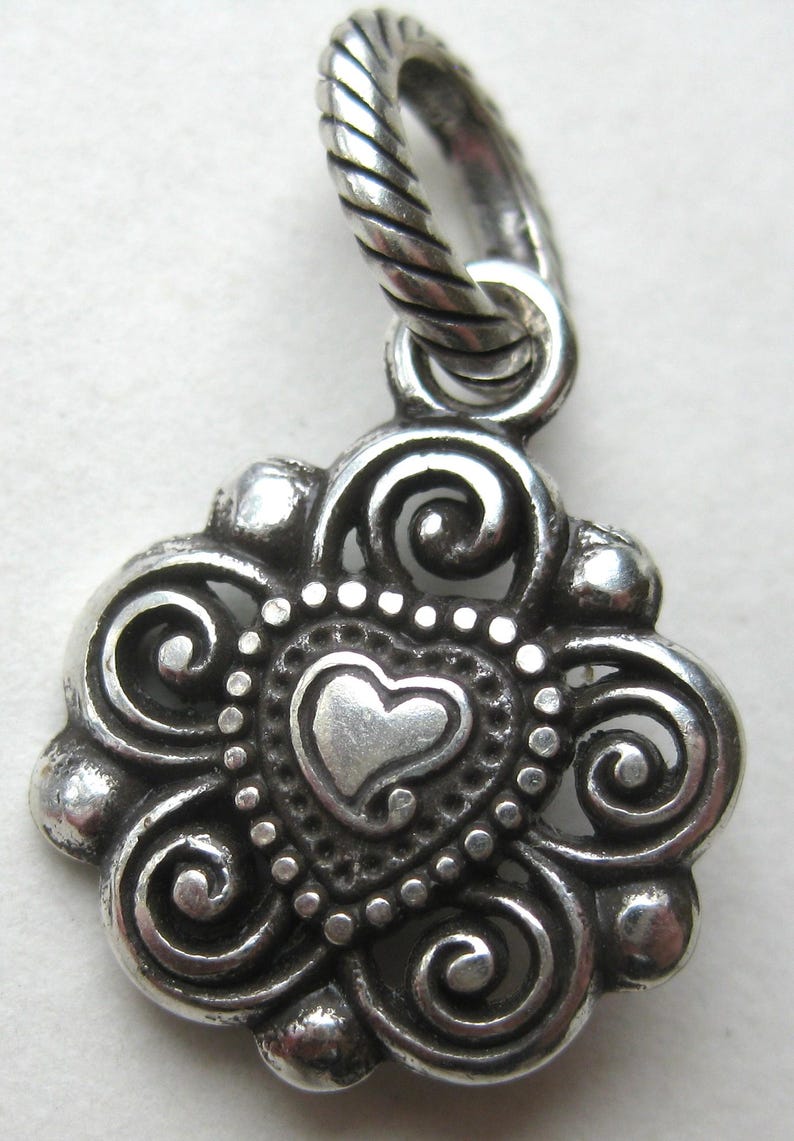 May include: A silver pendant featuring a central heart shape surrounded by intricate swirling metal patterns and a beaded border. The pendant is attached to a twisted rope-style silver bail at the top, showcasing a polished and oxidized finish.