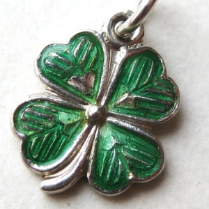 K&ouml;nnte beinhalten: Ein silberner Anh&auml;nger in Form eines vierbl&auml;ttrigen Kleeblatts. Das Kleeblatt hat leuchtend gr&uuml;ne Emaillebl&auml;tter mit detaillierten Adern, die von einem silbernen Rand umgeben sind. Es h&auml;ngt an einer kleinen Silberschlaufe.
