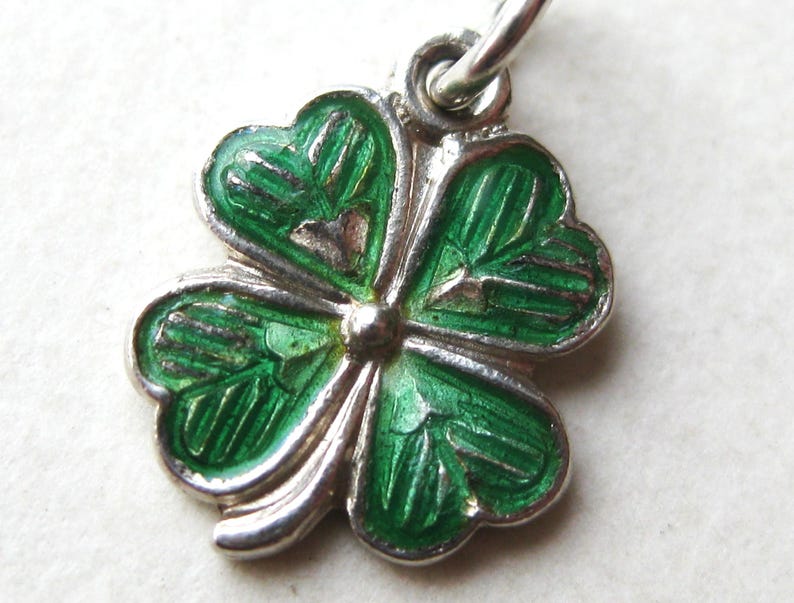 K&ouml;nnte beinhalten: Ein silberner Anh&auml;nger in Form eines vierbl&auml;ttrigen Kleeblatts. Das Kleeblatt hat leuchtend gr&uuml;ne Emaillebl&auml;tter mit detaillierten Adern, die von einem silbernen Rand umgeben sind. Es h&auml;ngt an einer kleinen Silberschlaufe.