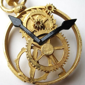 May include: A gold-toned metal pocket watch pendant featuring an open skeleton design that exposes the intricate internal gear mechanism. The piece includes two prominent black hands positioned against the textured gold gears and a circular frame.