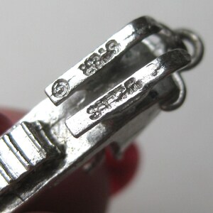 Może przedstawiać: Zbliżenie srebrnego zapięcia do biżuterii. Zapięcie ma prostokątny kształt z zawiasowym designem i małą pętlą. Metal jest wybity słowem "STERLING" i numerem "925".