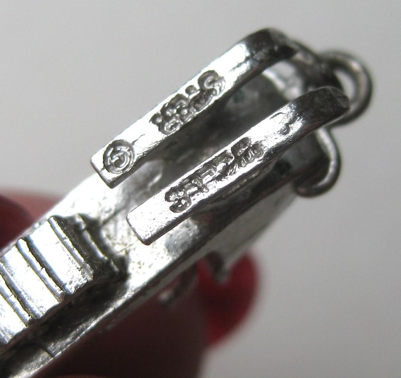 Może przedstawiać: Zbliżenie srebrnego zapięcia do biżuterii. Zapięcie ma prostokątny kształt z zawiasowym designem i małą pętlą. Metal jest wybity słowem "STERLING" i numerem "925".