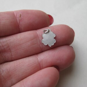 K&ouml;nnte beinhalten: Ein kleiner, silberfarbener Kleeblatt-Anh&auml;nger mit einer Schlaufe oben zum Befestigen an einer Halskette oder einem Armband. Der Anh&auml;nger wird zwischen den Fingern gehalten und zeigt sein filigranes Design.