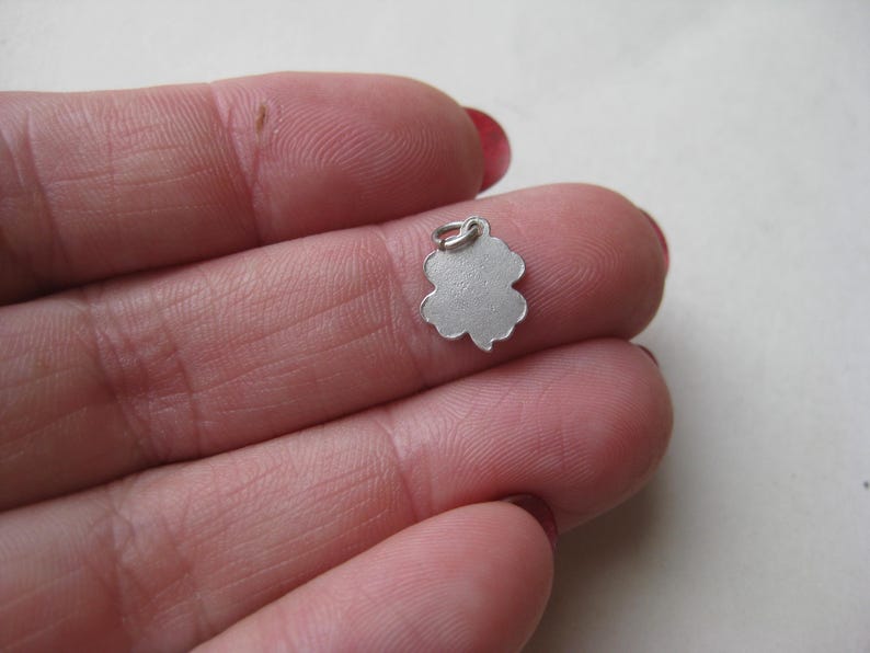 K&ouml;nnte beinhalten: Ein kleiner, silberfarbener Kleeblatt-Anh&auml;nger mit einer Schlaufe oben zum Befestigen an einer Halskette oder einem Armband. Der Anh&auml;nger wird zwischen den Fingern gehalten und zeigt sein filigranes Design.