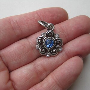 May include: A silver-toned metal pendant featuring a central heart-shaped blue gemstone surrounded by swirling metal patterns and small clear accent stones. The pendant includes a twisted metal bail at the top for attachment to a necklace chain.