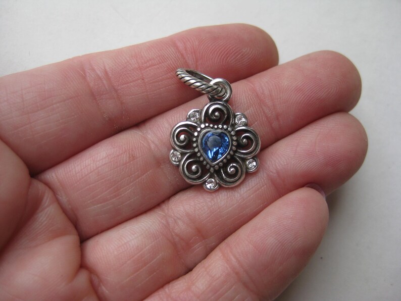 May include: A silver-toned metal pendant featuring a central heart-shaped blue gemstone surrounded by swirling metal patterns and small clear accent stones. The pendant includes a twisted metal bail at the top for attachment to a necklace chain.