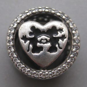 May include: A circular silver charm bead featuring a central heart-shaped cutout with an intricate filigree pattern. The outer ring is encrusted with small, clear, sparkling stones, creating a textured border around the dark, recessed heart center.