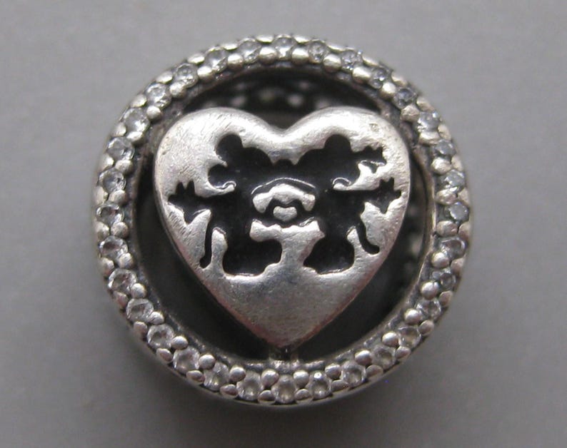 May include: A circular silver charm bead featuring a central heart-shaped cutout with an intricate filigree pattern. The outer ring is encrusted with small, clear, sparkling stones, creating a textured border around the dark, recessed heart center.