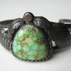 May include: A silver cuff bracelet featuring a central triangular green turquoise stone with dark matrix veining. The stone is framed by a twisted silver wire border and flanked by oxidised silver leaf motifs on both sides of the band.