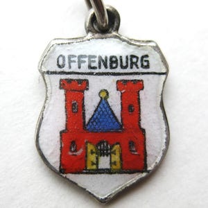 May include: A shield-shaped metal charm featuring a white enamel background with the text OFFENBURG in black letters. The design depicts a red castle with two towers, a blue roof, and open yellow gates, all enclosed within a silver-toned metal border.