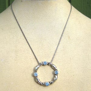 May include: Silver-toned necklace featuring a circular pendant with a braided design and six light blue gemstones. The pendant hangs from a delicate chain, creating a simple yet elegant accessory. The necklace is displayed on a mannequin.