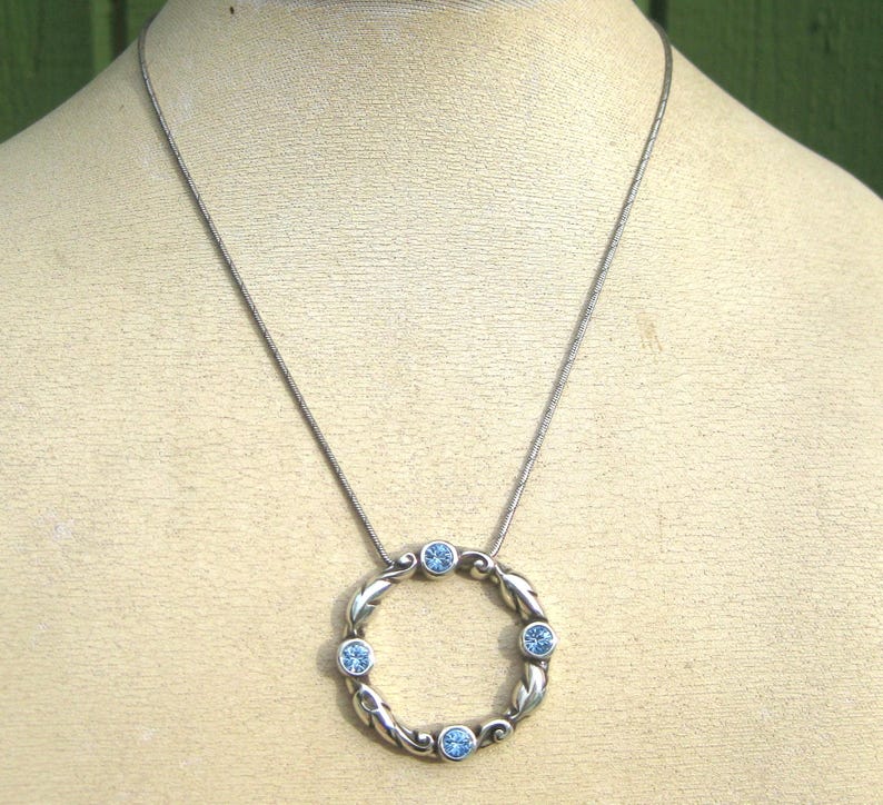 May include: Silver-toned necklace featuring a circular pendant with a braided design and six light blue gemstones. The pendant hangs from a delicate chain, creating a simple yet elegant accessory. The necklace is displayed on a mannequin.