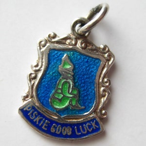 Könnte beinhalten: Ein silberfarbener Anhänger mit blauem und grünem Emaille-Design. Der Anhänger hat die Form eines Schildes mit einer sitzenden Figur und den Worten "Piskie Good Luck" in blauen Buchstaben. Der Anhänger hat einen dekorativen Rand und eine Schlaufe zur Befestigung.