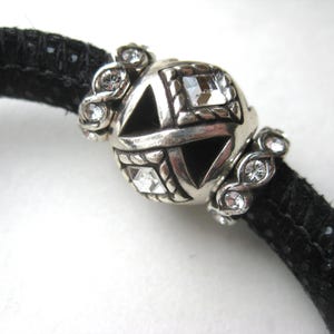 Puede incluir: Una pulsera de cord&oacute;n negro texturizado con una cuenta central esf&eacute;rica de tono plateado con recortes geom&eacute;tricos y cristales cuadrados transparentes, flanqueada por dos cuentas espaciadoras plateadas con peque&ntilde;os cristales redondos.