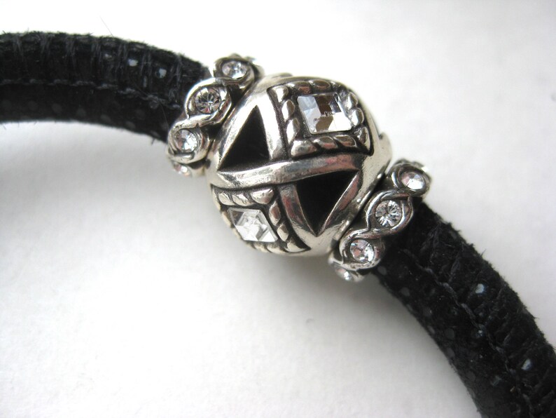 Puede incluir: Una pulsera de cord&oacute;n negro texturizado con una cuenta central esf&eacute;rica de tono plateado con recortes geom&eacute;tricos y cristales cuadrados transparentes, flanqueada por dos cuentas espaciadoras plateadas con peque&ntilde;os cristales redondos.