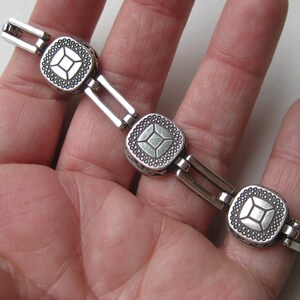 May include: A silver-toned metal link bracelet featuring three square-shaped charms. Each charm has a central square design surrounded by a textured border of small raised dots. The links connecting the charms are rectangular and open in the center.