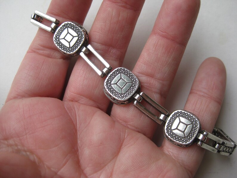 May include: A silver-toned metal link bracelet featuring three square-shaped charms. Each charm has a central square design surrounded by a textured border of small raised dots. The links connecting the charms are rectangular and open in the center.