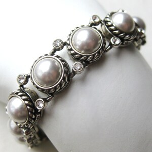 Puede incluir: Una pulsera de eslabones en tono plateado con perlas blancas redondas engastadas en bordes de metal trenzado. Peque&ntilde;os cristales redondos transparentes y facetados se sit&uacute;an entre cada perla, creando un patr&oacute;n repetitivo a lo largo de la pieza.