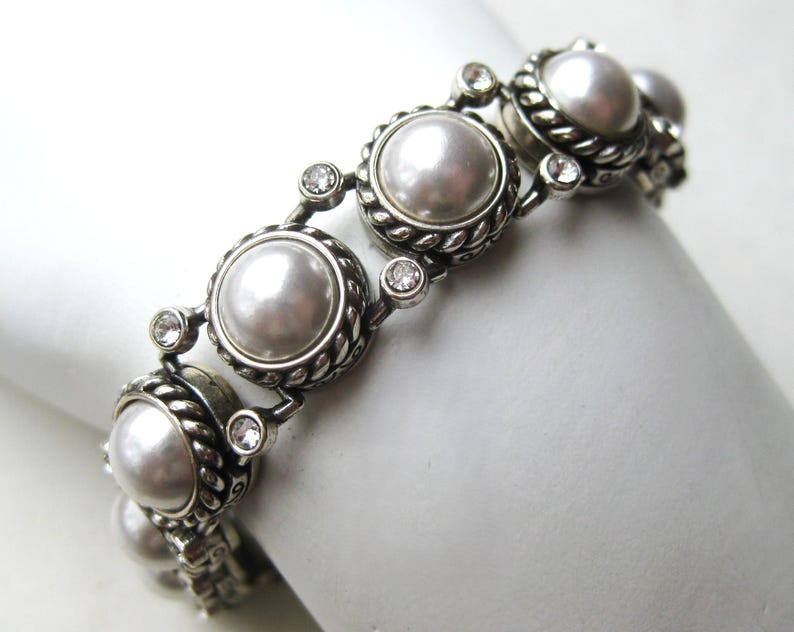 Puede incluir: Una pulsera de eslabones en tono plateado con perlas blancas redondas engastadas en bordes de metal trenzado. Peque&ntilde;os cristales redondos transparentes y facetados se sit&uacute;an entre cada perla, creando un patr&oacute;n repetitivo a lo largo de la pieza.