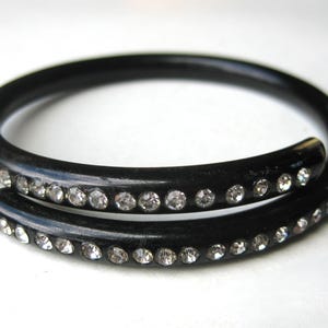 May include: A black, spiral-shaped bracelet with a row of clear, sparkling rhinestones. The bracelet is made of a dark, possibly lacquered material, and the rhinestones are evenly spaced along the surface.