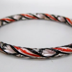 May include: A clear glass bangle bracelet with a twisted design. The bracelet features red, black, and white stripes that wrap around the circular form. The bracelet is a piece of jewelry.