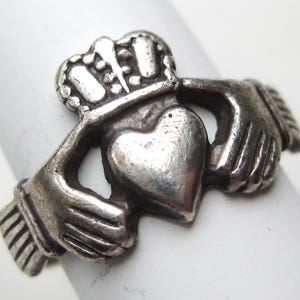 May include: A silver Claddagh ring featuring a heart held by two hands, topped with a crown. The ring is worn on a white surface, showcasing the intricate details of the design. This ring is a symbol of love, loyalty, and friendship.