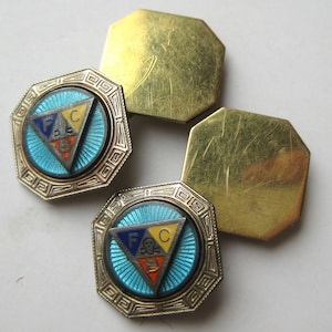 May include: Vintage cufflinks with a geometric design. The cufflinks feature a light blue enamel center with a triangle containing the letters F, A, and C. The outer frame is a silver color with a gold-colored back.