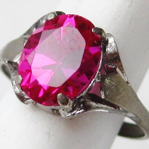May include: A silver-toned ring featuring a large, oval-cut, hot pink gemstone. The gemstone is the focal point, set in a pronged setting. The ring's band has a simple, slightly textured design.