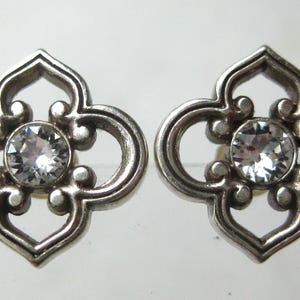 May include: A pair of silver-toned metal stud earrings featuring a four-lobed openwork design with a central clear, round-cut faceted stone. The metal has a polished finish with rounded edges, creating a symmetrical, decorative floral-like shape.