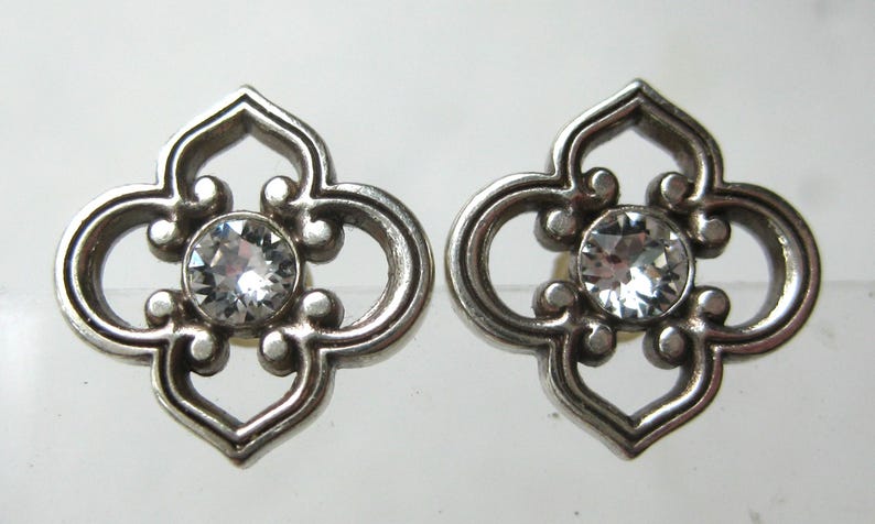 May include: A pair of silver-toned metal stud earrings featuring a four-lobed openwork design with a central clear, round-cut faceted stone. The metal has a polished finish with rounded edges, creating a symmetrical, decorative floral-like shape.