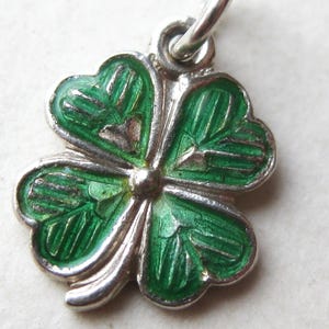 K&ouml;nnte beinhalten: Ein silberfarbener vierbl&auml;ttriger Kleeblatt-Anh&auml;nger mit gr&uuml;nen Emaille-Details. Der Anh&auml;nger hat eine kleine Schlaufe zum Befestigen an einer Halskette oder einem Armband. Die Bl&auml;tter des Kleeblatts haben ein detailliertes, strukturiertes Aussehen.