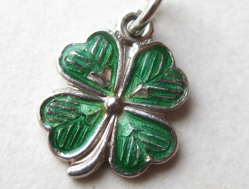 K&ouml;nnte beinhalten: Ein silberfarbener vierbl&auml;ttriger Kleeblatt-Anh&auml;nger mit gr&uuml;nen Emaille-Details. Der Anh&auml;nger hat eine kleine Schlaufe zum Befestigen an einer Halskette oder einem Armband. Die Bl&auml;tter des Kleeblatts haben ein detailliertes, strukturiertes Aussehen.