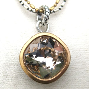 Puede incluir: Un collar con colgante que presenta un cristal facetado de color champ&aacute;n y forma cuadrada, engastado en un bisel dorado mate. El colgante cuelga de una anilla plateada trenzada unida a una cadena de dos hilos con cuentas plateadas y doradas.
