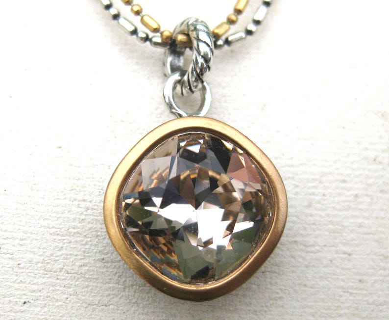 Puede incluir: Un collar con colgante que presenta un cristal facetado de color champ&aacute;n y forma cuadrada, engastado en un bisel dorado mate. El colgante cuelga de una anilla plateada trenzada unida a una cadena de dos hilos con cuentas plateadas y doradas.