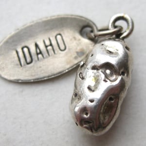 May include: A silver-toned metal charm featuring a textured, oblong potato shape alongside an oval-shaped tag stamped with the word IDAHO in capital letters. Both pieces are connected by a small circular jump ring at the top of the charm.