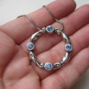 May include: A silver-toned pendant necklace featuring a circular design with six light blue gemstones. The pendant has a decorative, flowing pattern. The necklace is displayed on a silver chain.