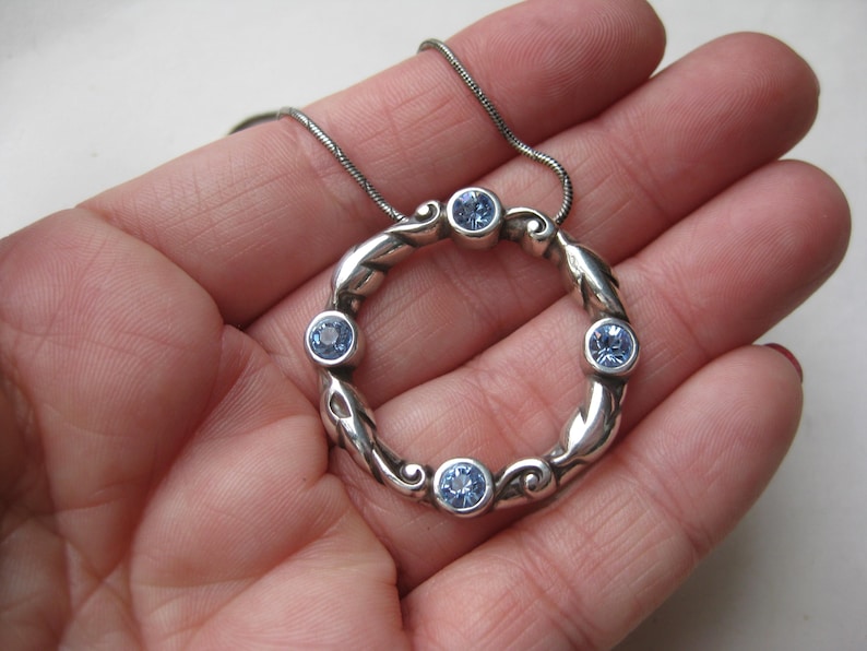 May include: A silver-toned pendant necklace featuring a circular design with six light blue gemstones. The pendant has a decorative, flowing pattern. The necklace is displayed on a silver chain.