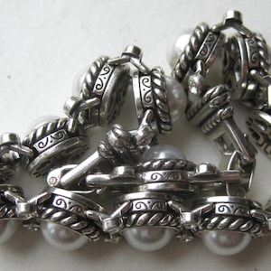 Puede incluir: Una pulsera de eslabones plateados con perlas blancas redondas de imitaci&oacute;n engastadas en monturas met&aacute;licas texturizadas y ornamentadas. Los eslabones est&aacute;n unidos por bordes met&aacute;licos trenzados y patrones de volutas, con un cierre met&aacute;lico.