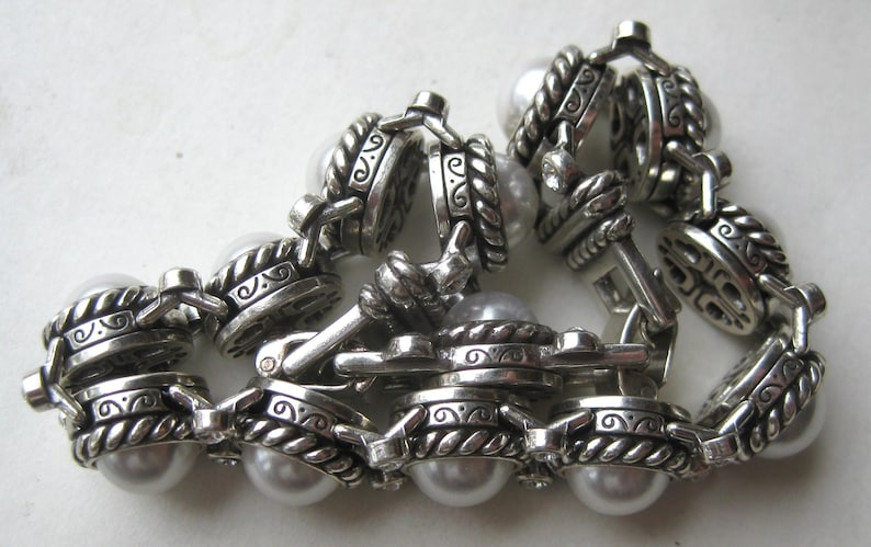 Puede incluir: Una pulsera de eslabones plateados con perlas blancas redondas de imitaci&oacute;n engastadas en monturas met&aacute;licas texturizadas y ornamentadas. Los eslabones est&aacute;n unidos por bordes met&aacute;licos trenzados y patrones de volutas, con un cierre met&aacute;lico.