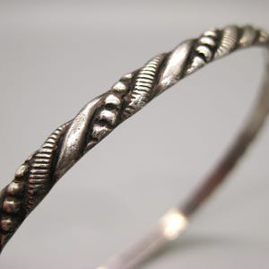 May include: A silver-toned metal bangle bracelet featuring a twisted rope design. The surface of the bracelet displays a textured pattern with alternating smooth, curved segments and small, raised bead-like details along the entire length.