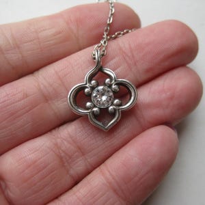May include: A silver-toned pendant necklace featuring a four-lobed, openwork metal frame with a central, clear, round-cut gemstone. The pendant is attached to a delicate, matching silver-toned metal chain link necklace.