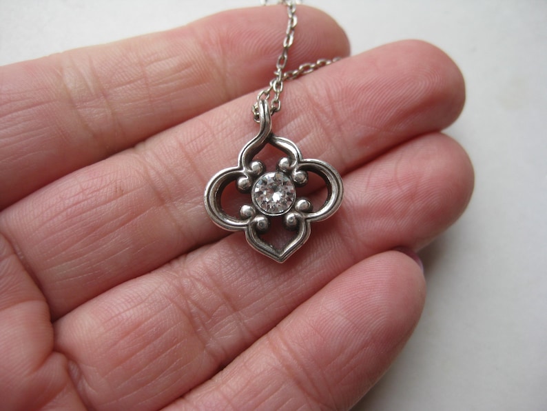 May include: A silver-toned pendant necklace featuring a four-lobed, openwork metal frame with a central, clear, round-cut gemstone. The pendant is attached to a delicate, matching silver-toned metal chain link necklace.