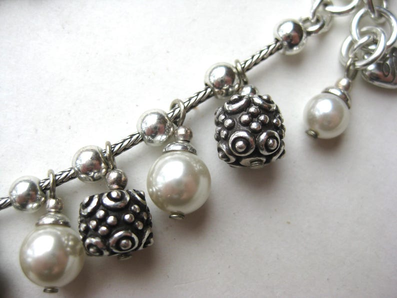 Puede incluir: Primer plano de una pulsera de abalorios plateada con abalorios decorativos. La pulsera presenta una cadena trenzada y varios abalorios, incluyendo perlas de imitaci&oacute;n y cuentas texturizadas con dise&ntilde;os. Los abalorios cuelgan de la cadena.